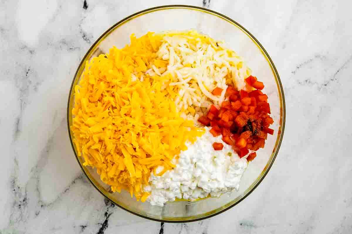 A glass bowl contains shredded cheddar and mozzarella cheese, cottage cheese, chopped red bell pepper, and seasonings on a marble countertop.