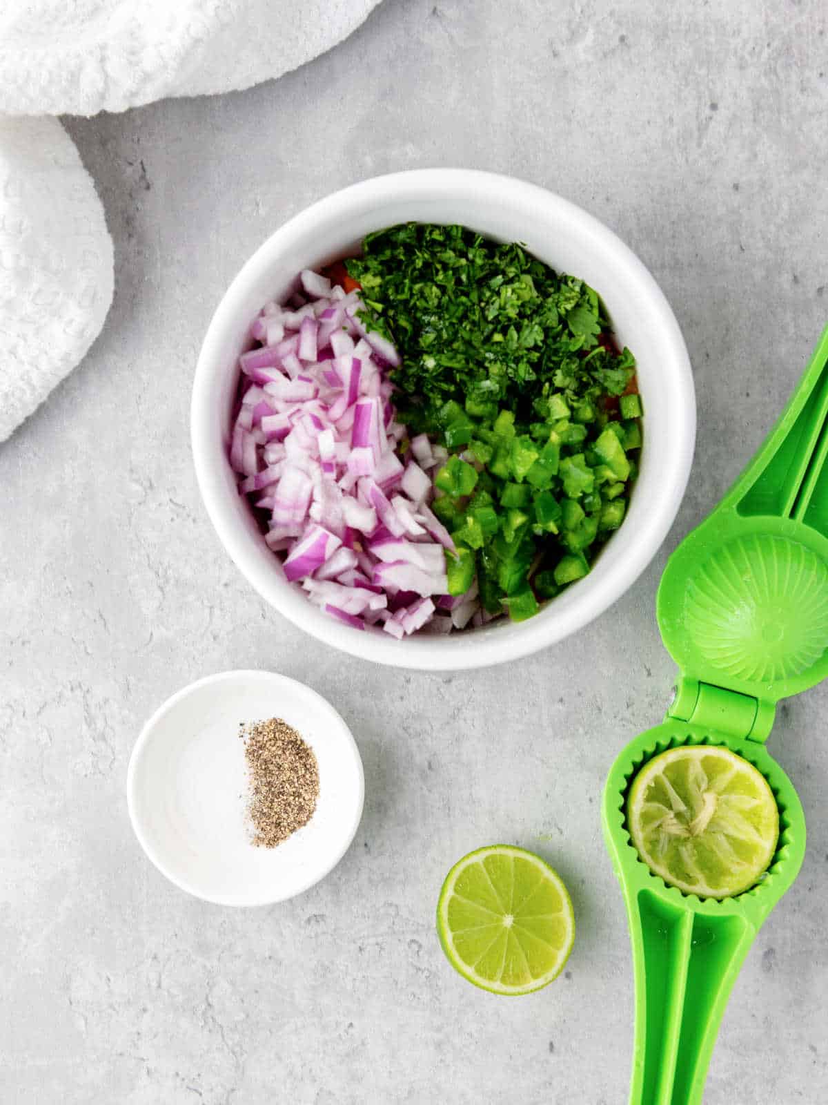 Overhead view of a bowl with chopped red onion, green pepper, and cilantro, next to a dish of black pepper and a lime squeezer with squeezed lime halves and a halved lime.