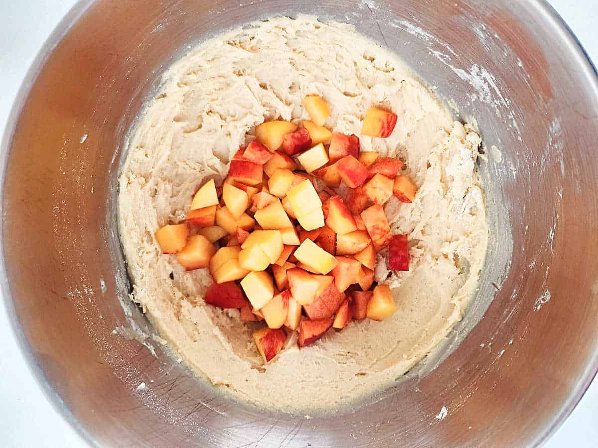 A stainless steel bowl containing cookie dough with chopped peaches on top, ready to be mixed together.