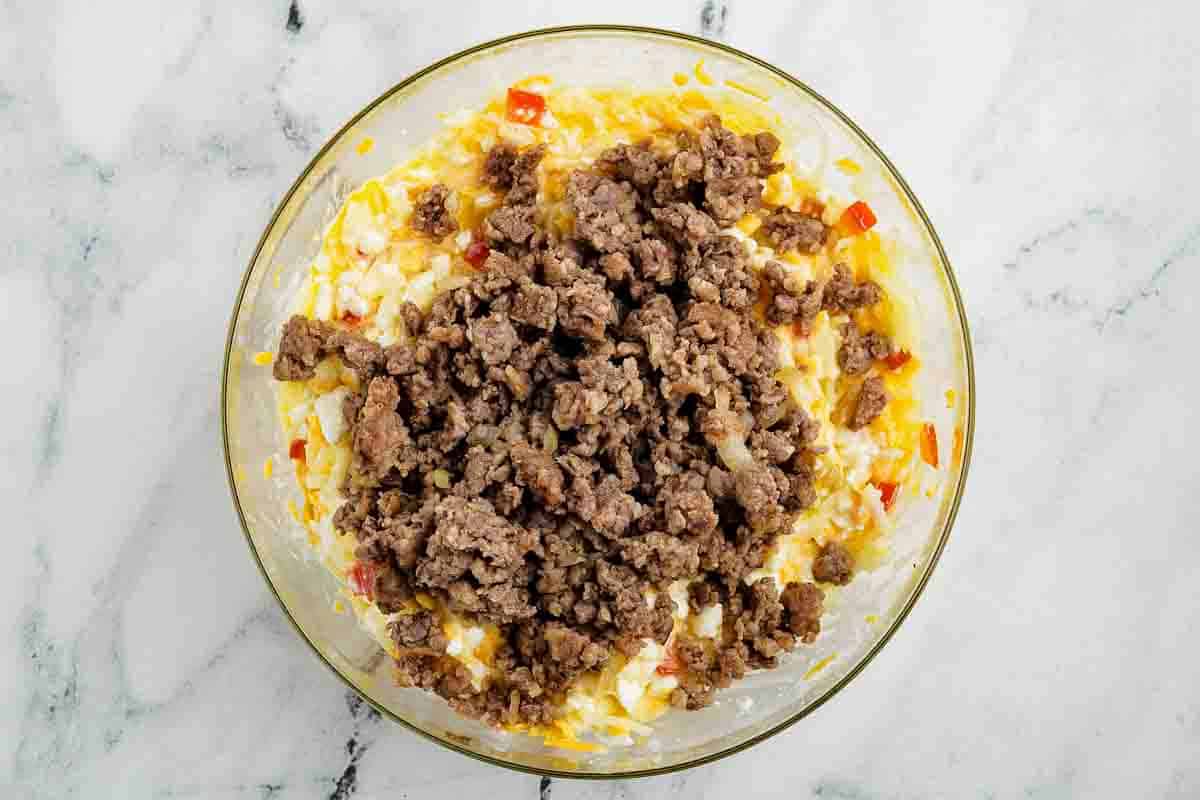 A glass bowl containing cooked ground sausage on top of a mixture of chopped vegetables, cheese, and eggs, placed on a marble surface.