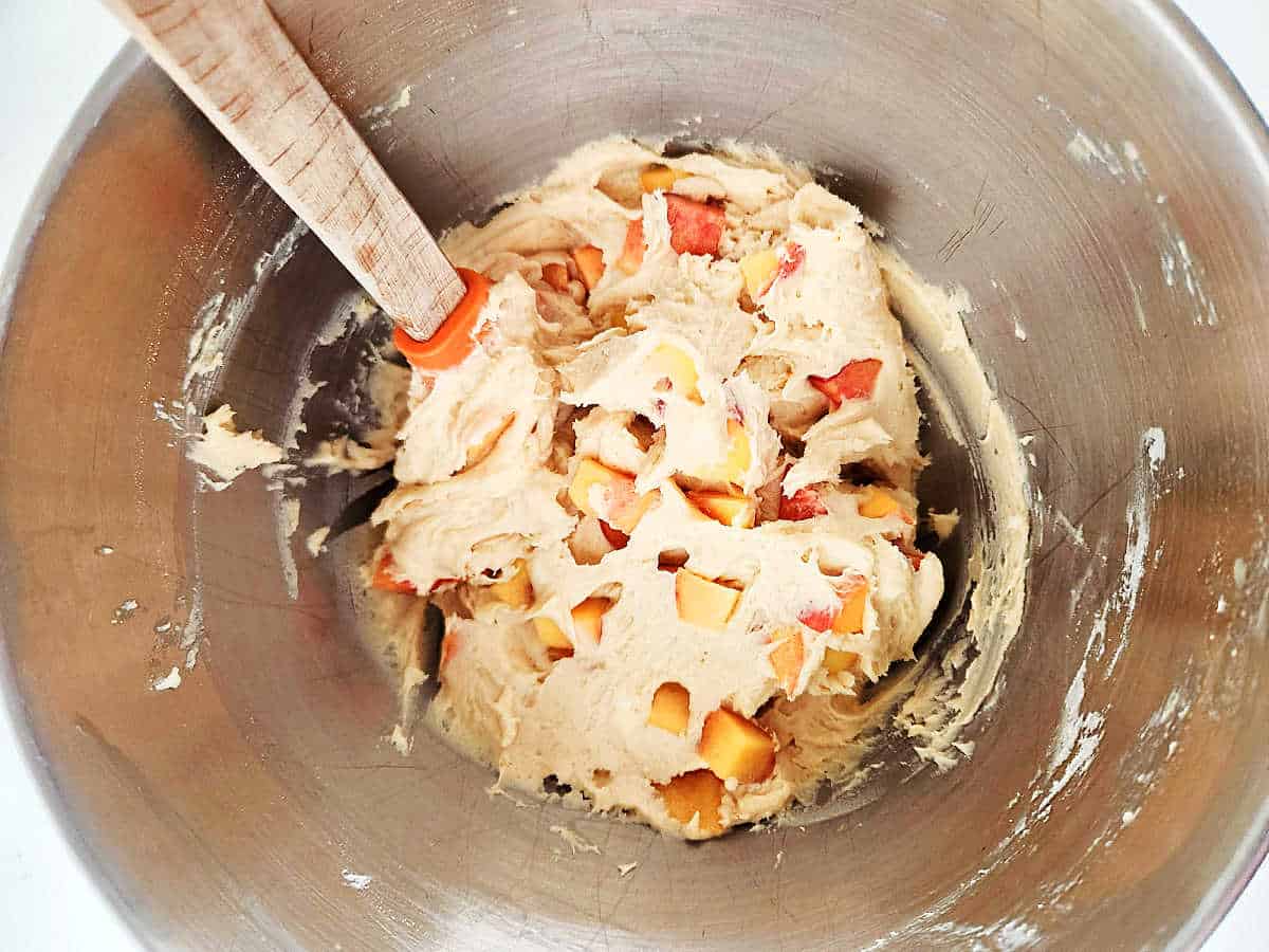 Stainless steel bowl with creamy batter mixed with chopped fruit, being stirred by a spatula.