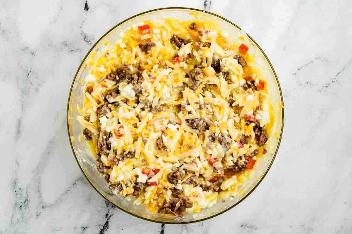 A glass bowl filled with a mixture of shredded cheese, cooked ground meat, diced red peppers, and other ingredients on a marble surface.
