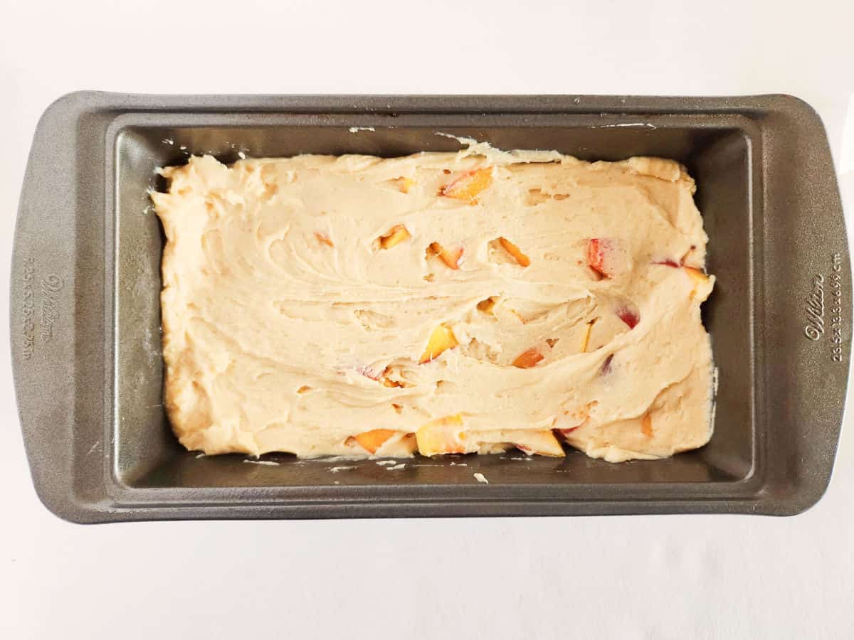 Rectangular metal loaf pan filled with unbaked batter and visible peach chunks, placed on a white surface.