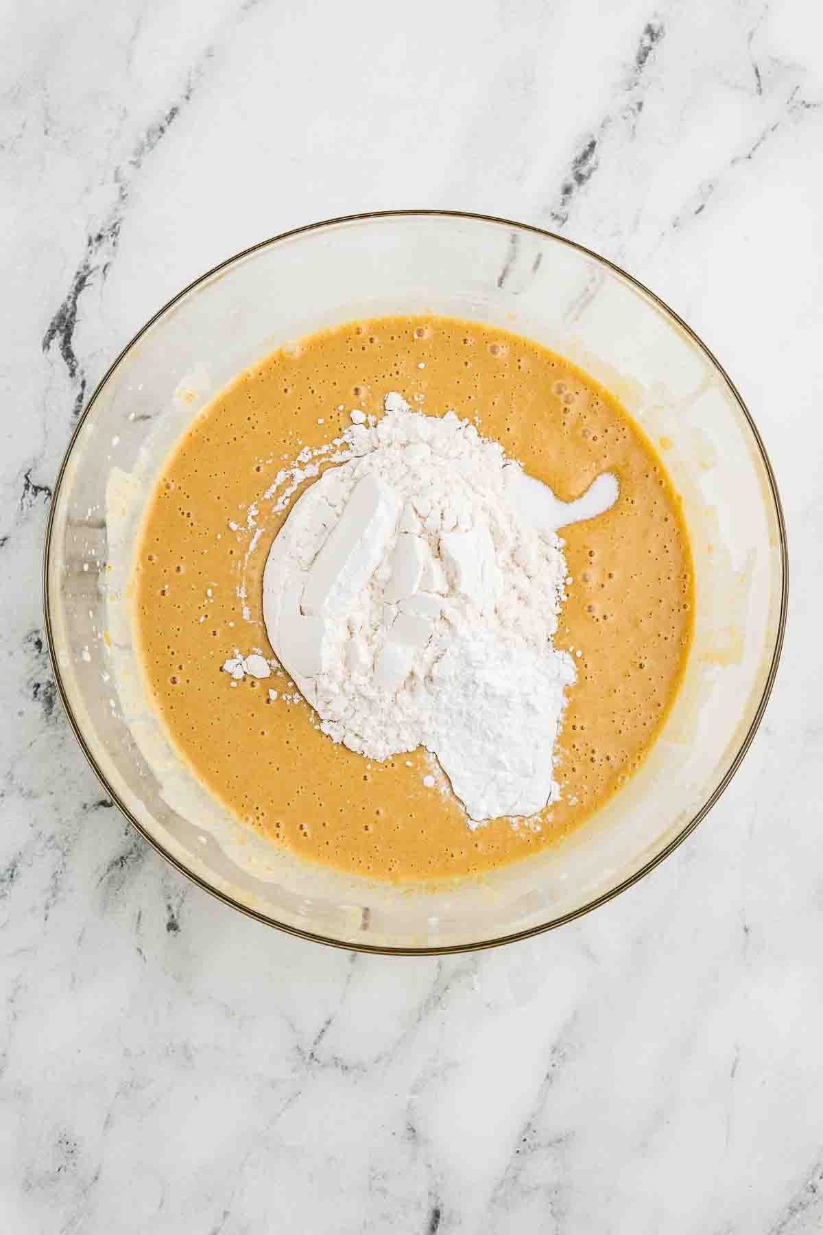 A glass bowl with a yellowish batter, topped with white flour and baking powder, sits on a white marble surface.