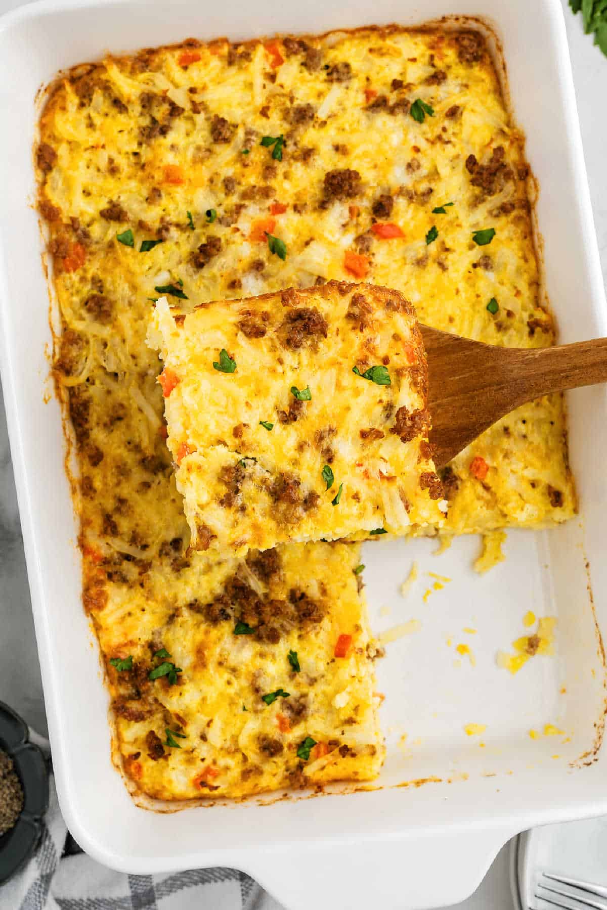 A breakfast casserole in a white baking dish with a wooden spatula lifting a square piece; eggs, sausage, cheese, and vegetables are visible.