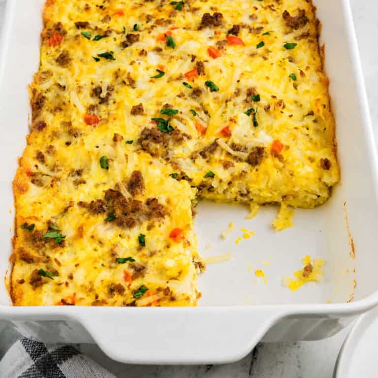 Baked breakfast casserole with hash browns, ground meat, egg, and vegetables is served in a white dish with a square piece removed.