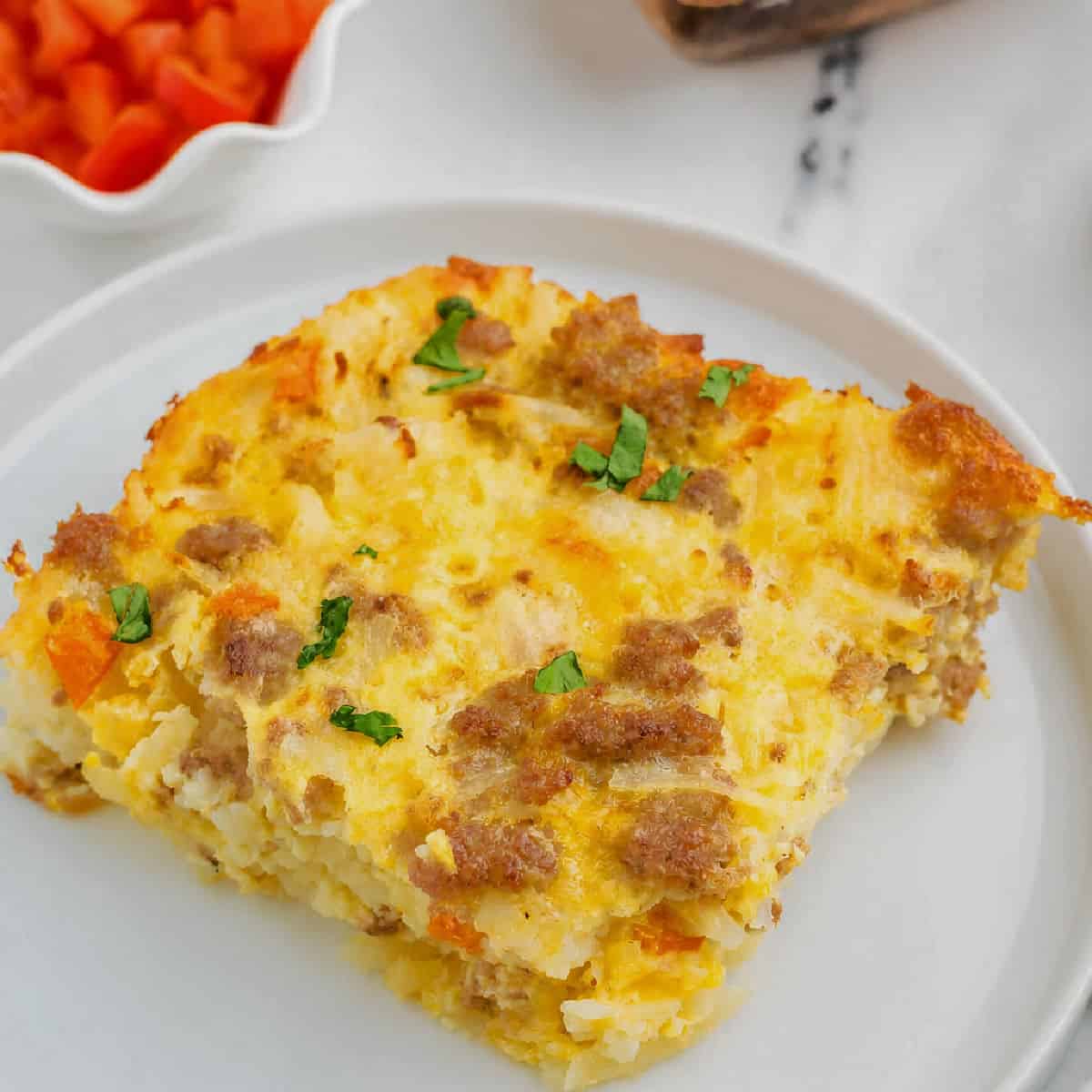 A slice of baked breakfast casserole with sausage, cheese, and herbs sits on a white plate, with diced red peppers in the background.