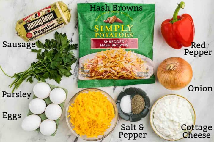 Ingredients for a breakfast casserole: sausage, hash browns, red pepper, onion, parsley, eggs, shredded cheese, salt, pepper, and cottage cheese arranged on a white surface.