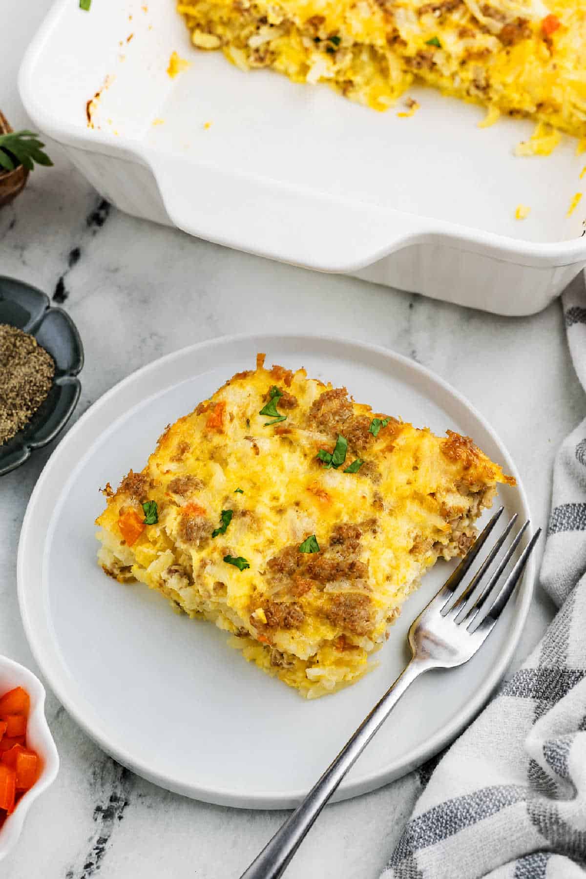 A square slice of egg, sausage breakfast casserole with vegetables and sausage on a plate, next to a fork; the rest of the casserole is in a white baking dish in the background.