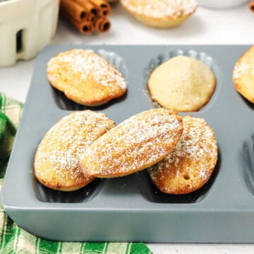 Madeleine cookies dusted with powdered sugar rest on and inside a nonstick madeleine baking tray, with a green checkered cloth nearby.