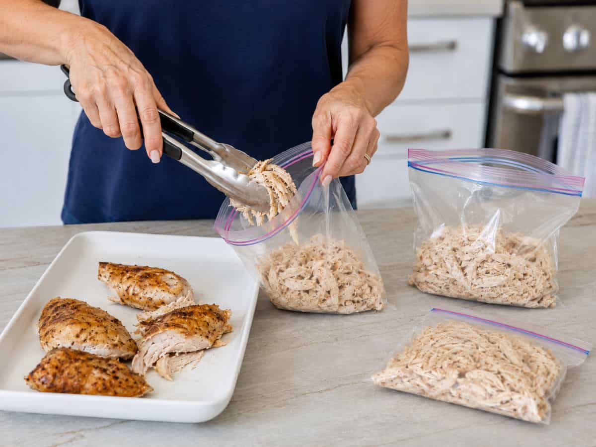 A person uses tongs to place shredded cooked chicken into a resealable bag, with more bags of shredded chicken and whole chicken breasts on the counter.