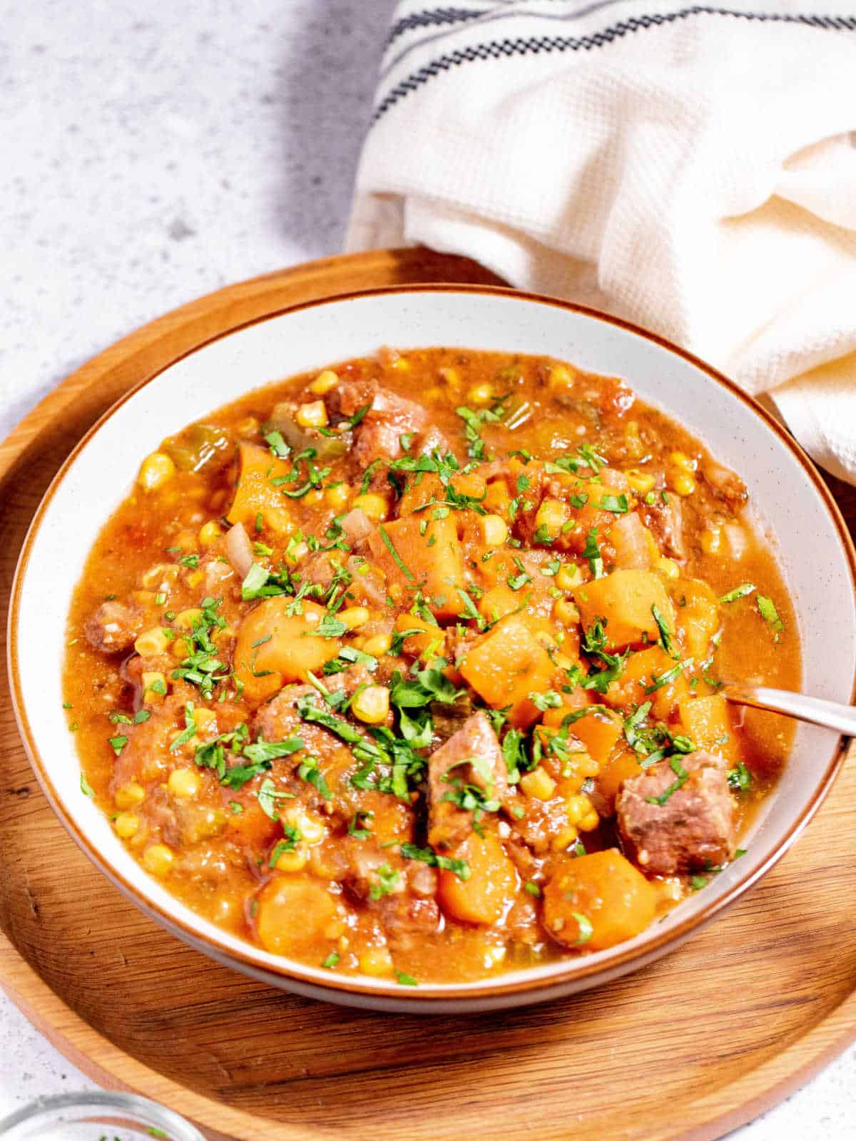 A bowl of hearty stew with chunks of beef, potatoes, corn, and vegetables, garnished with chopped herbs, served on a wooden tray with a spoon.