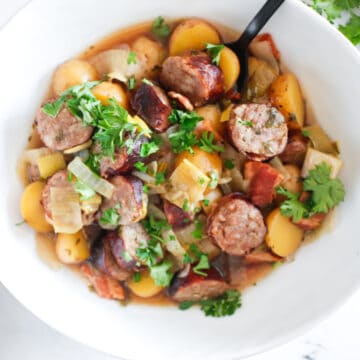 A bowl of Dublin coddle stew with sliced sausage, potatoes, onions, tomatoes, and fresh parsley, served with a black spoon.