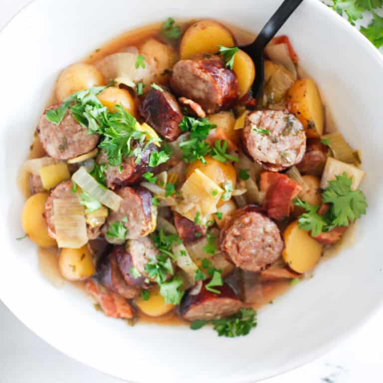 A bowl of Dublin coddle stew with sliced sausage, potatoes, onions, tomatoes, and fresh parsley, served with a black spoon.