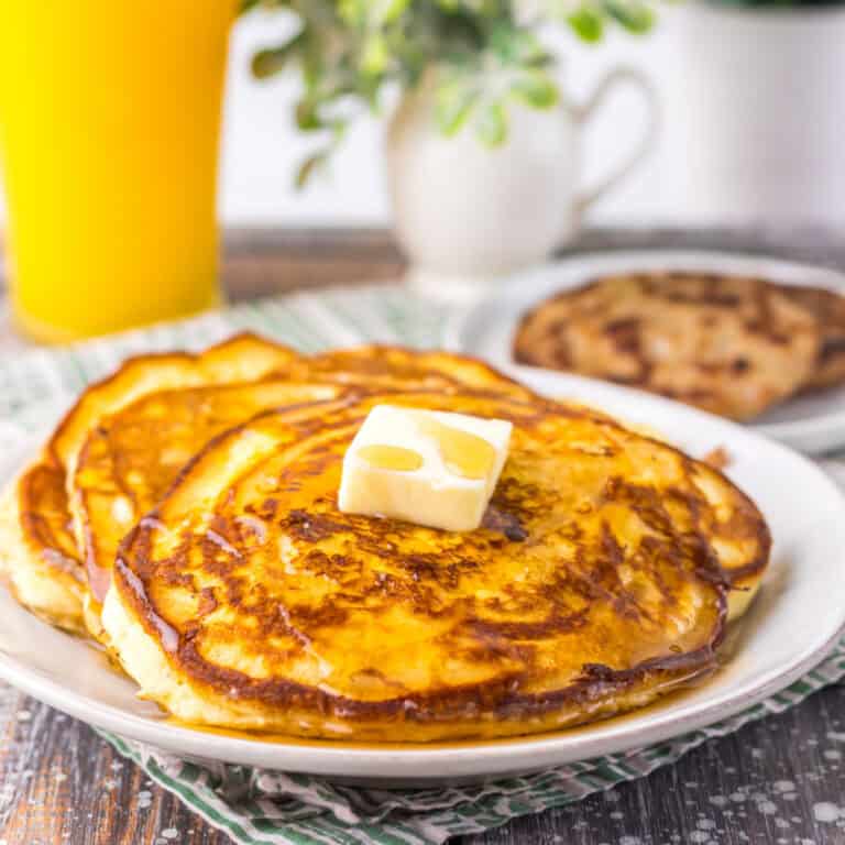 A plate of pancakes topped with a pat of melting butter, with a glass of orange juice and a plant in the background.