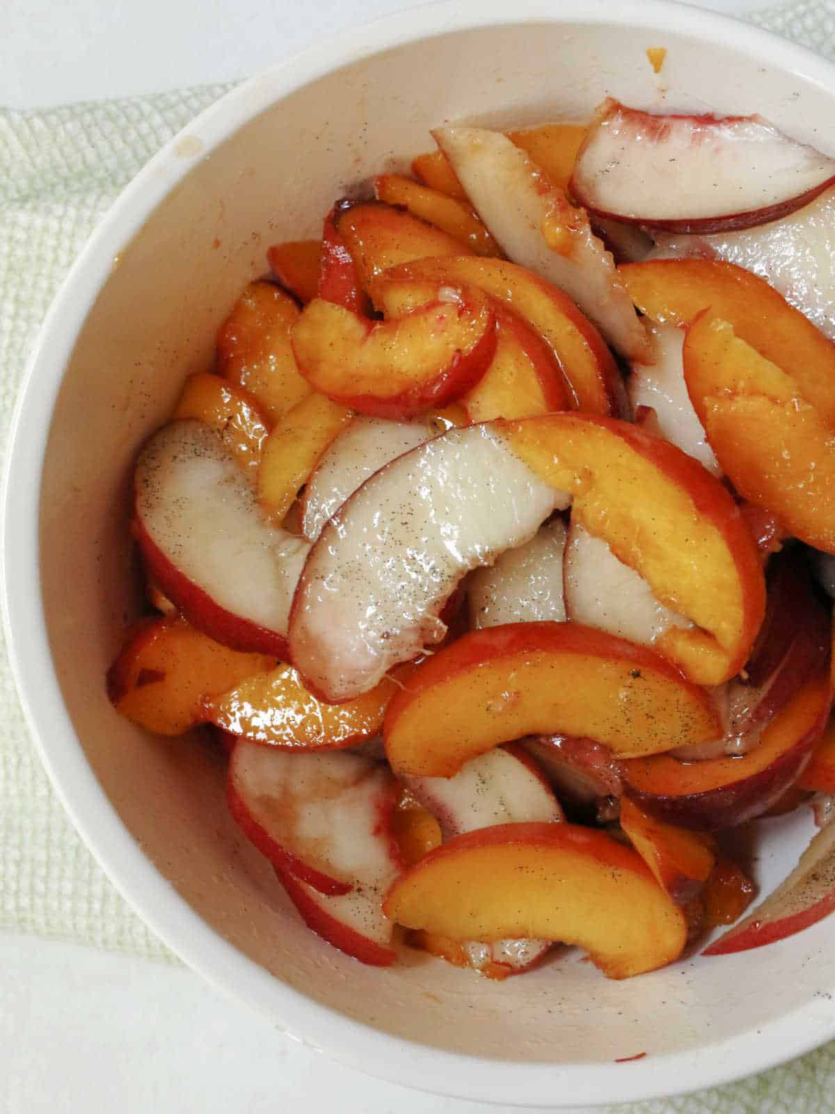 Sliced peaches and nectarines in a white bowl, sprinkled with spices, on a light green textured placemat.