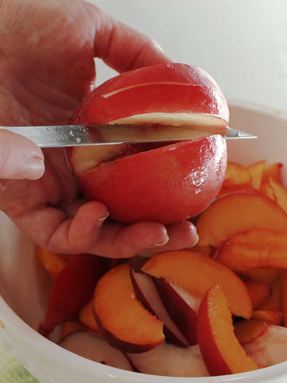 A hand holds a nectarine while slicing it with a knife over a bowl filled with sliced nectarines.