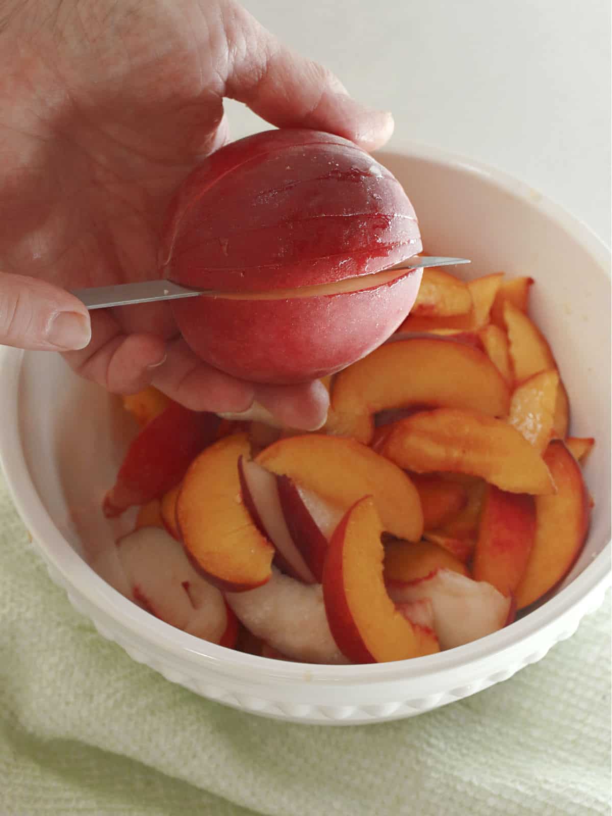 A hand slices a nectarine over a white bowl filled with nectarine wedges, with a green cloth beneath the bowl.