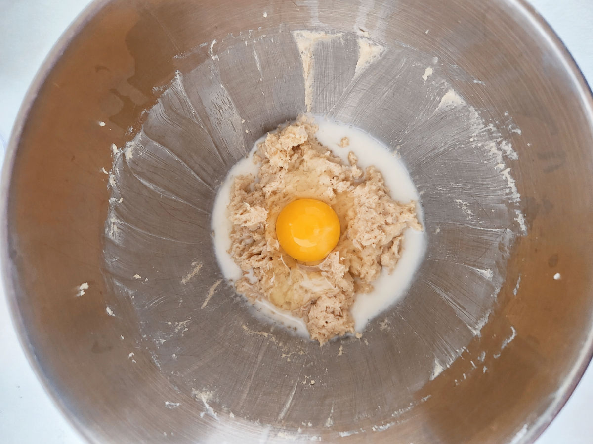 A stainless steel bowl contains dough, a cracked raw egg, and some milk in the center.
