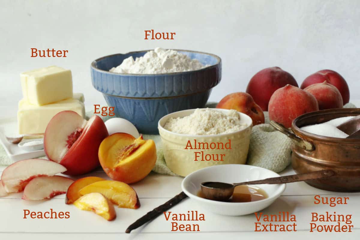 Arrangement of baking ingredients includes flour, almond flour, sugar, butter, eggs, peaches, vanilla bean, vanilla extract, and baking powder on a white surface with a bowl and towel.