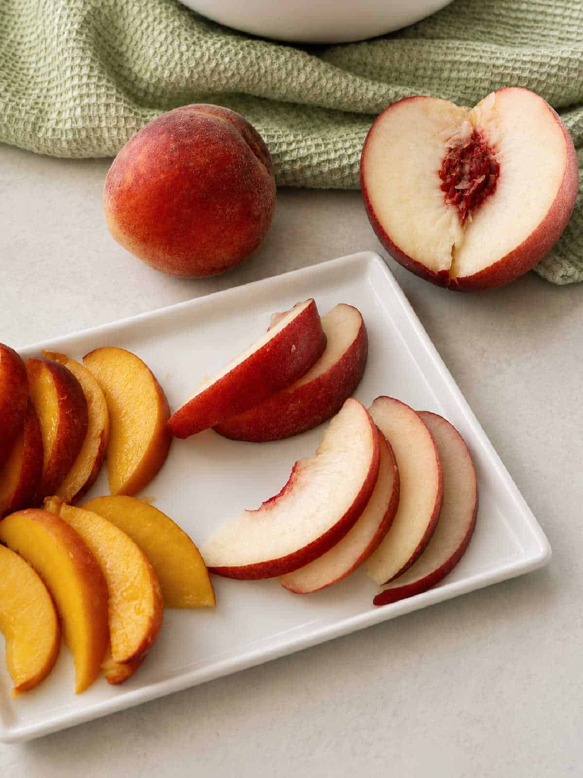 Whole and halved peaches rest on a countertop beside a white plate holding neatly arranged slices of yellow and white peaches.
