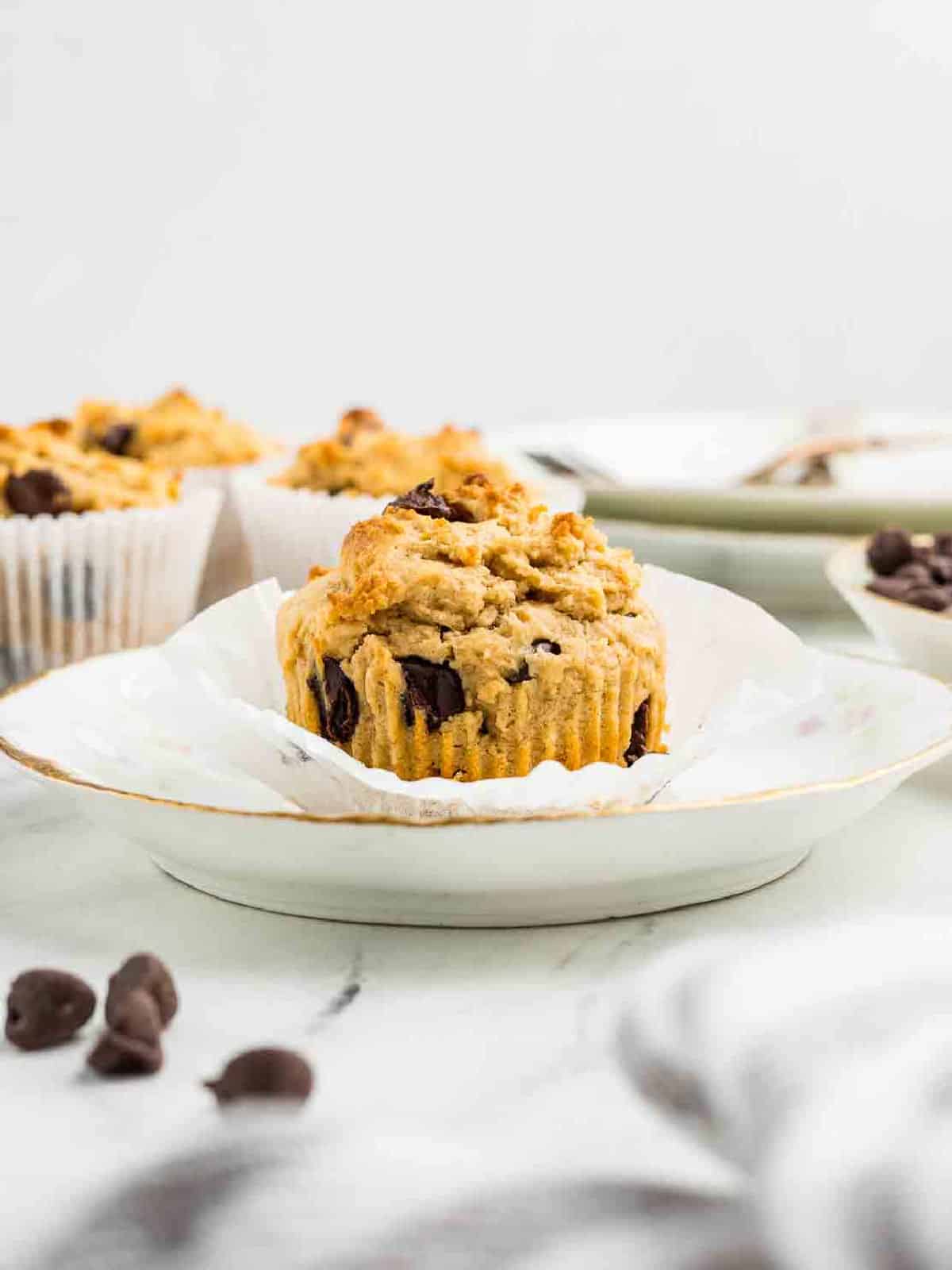 A chocolate chip muffin sits on a white plate, partially unwrapped from a paper liner, with more muffins and chocolate chips in the background.