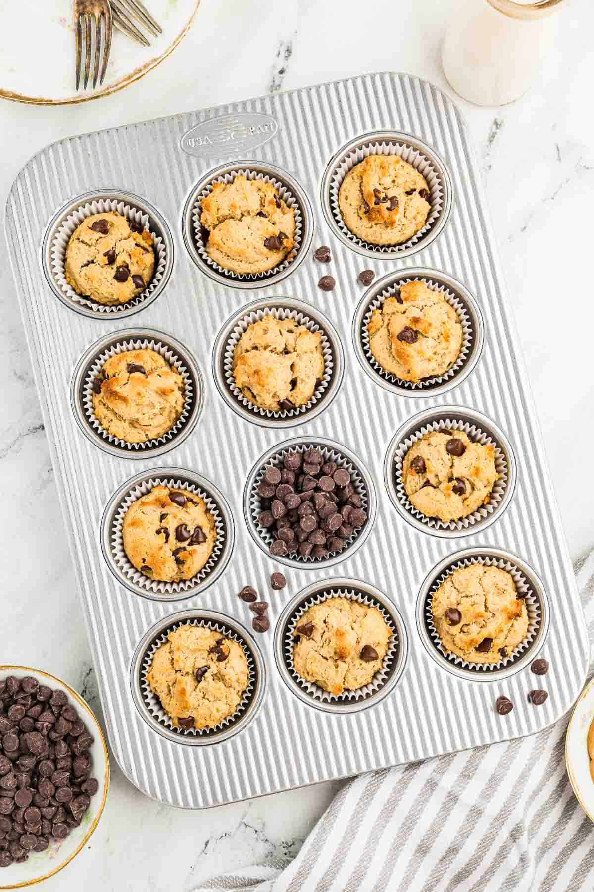 A muffin tin with nine unbaked chocolate chip muffins in paper liners and three cups filled with chocolate chips, on a marble surface.