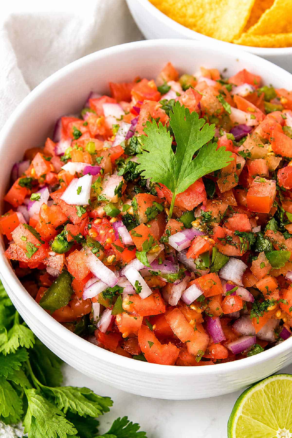A bowl of fresh pico de gallo salsa with chopped tomatoes, onions, cilantro, and jalapeños, garnished with a cilantro leaf. Lime and tortilla chips are nearby.