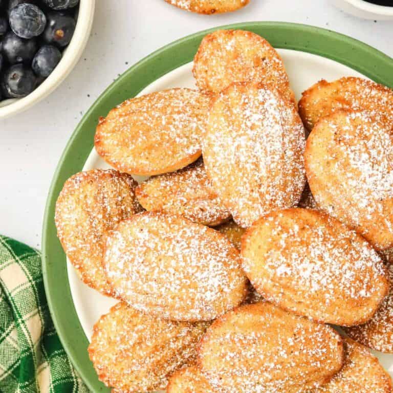 A plate of madeleines dusted with powdered sugar, next to a bowl of blueberries and a green checkered cloth.