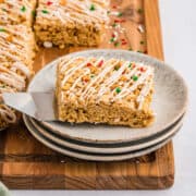 A rice krispie treat square with white drizzle and red and green sprinkles sits on a stack of plates, with a spatula underneath.