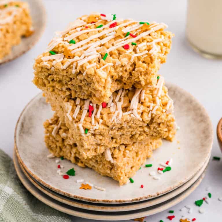 Three rice krispie treats stacked on a plate, topped with white drizzle and colorful sprinkles, with more treats and a glass of milk in the background.