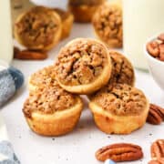 A stack of mini pecan tarts on a white surface, surrounded by whole pecans and a bowl of pecans in the background.