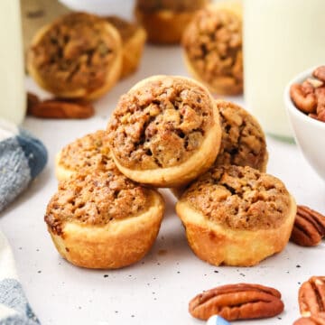A stack of mini pecan tarts on a white surface, surrounded by whole pecans and a bowl of pecans in the background.