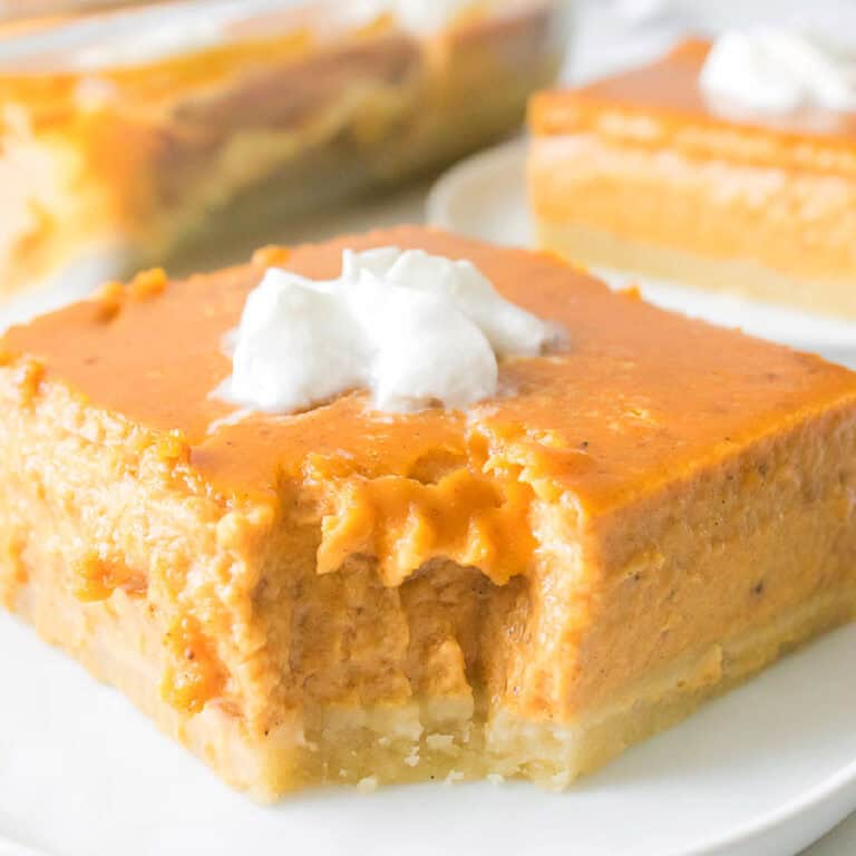 A square slice of pumpkin pie bar with a bite taken out, topped with whipped cream, sits on a white plate. More bars are visible in the background.