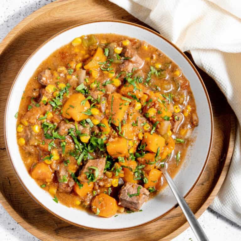 A bowl of beef stew with chunks of meat, potatoes, corn, carrots, and herbs, served on a white plate with a spoon.