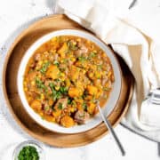 A bowl of hearty stew with chunks of meat, potatoes, carrots, and corn, garnished with fresh herbs, sits on a wooden plate with a spoon and a napkin.
