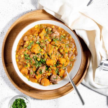 A bowl of hearty stew with chunks of meat, potatoes, carrots, and corn, garnished with fresh herbs, sits on a wooden plate with a spoon and a napkin.