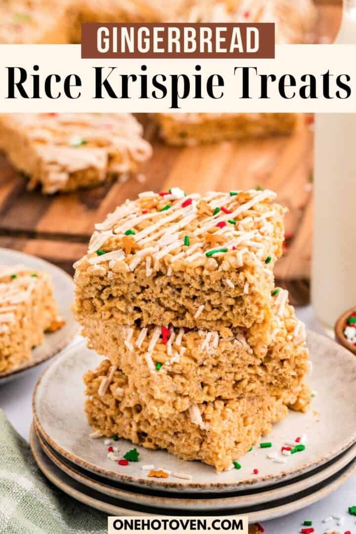 Three gingerbread Rice Krispie treats with white drizzle and festive sprinkles are stacked on a plate, with more treats and a glass of milk in the background.