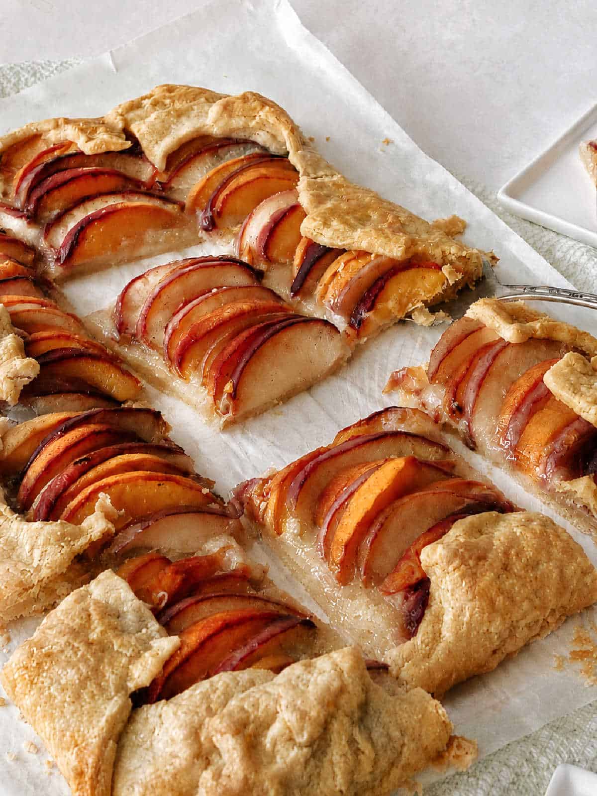 Rectangular fruit galette with golden crust, topped with neatly arranged slices of peaches and plums, cut into square pieces on parchment paper.
