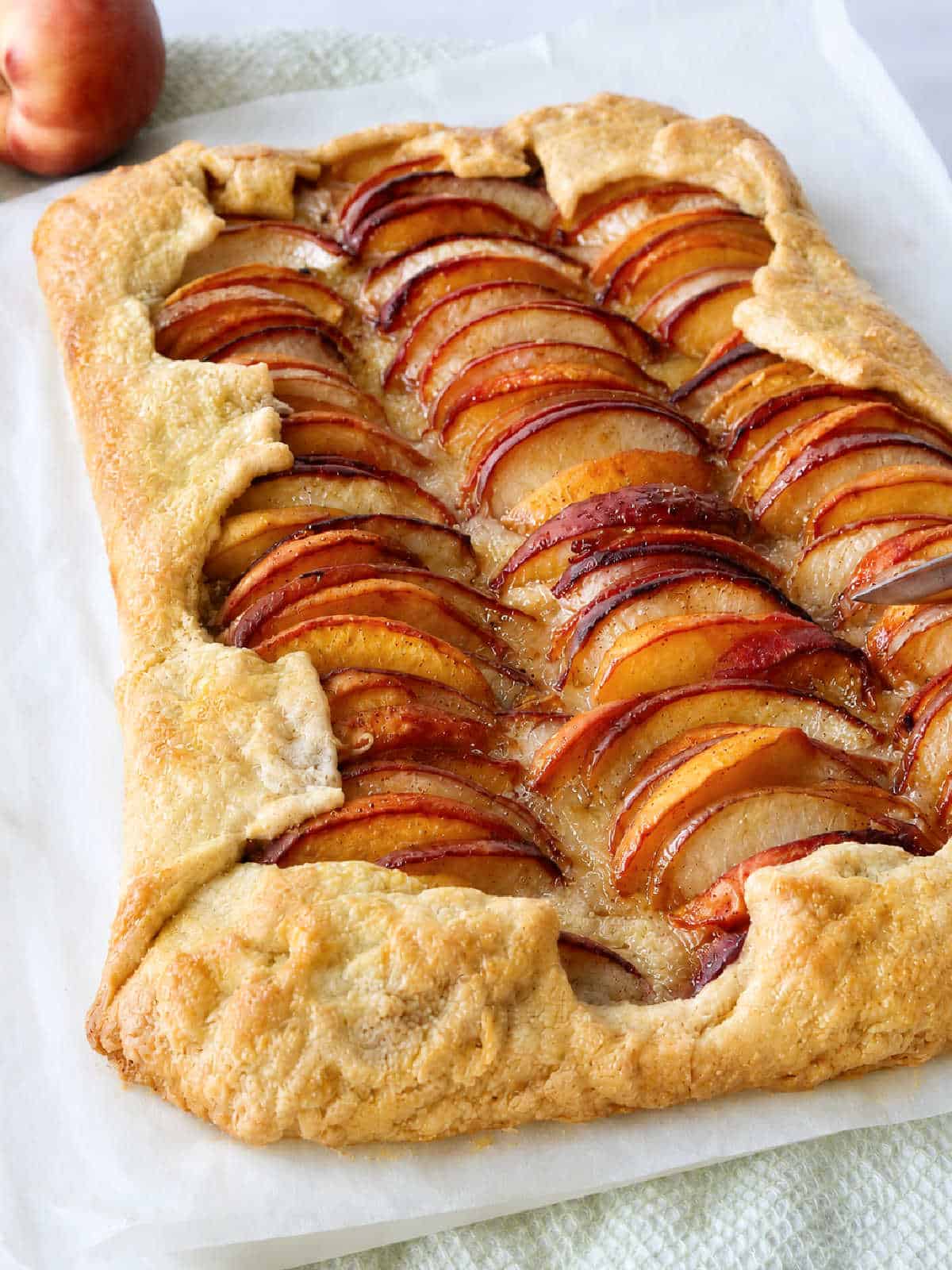 A rectangular fruit galette with golden, flaky crust, topped with neatly arranged slices of peach, sits on parchment paper.