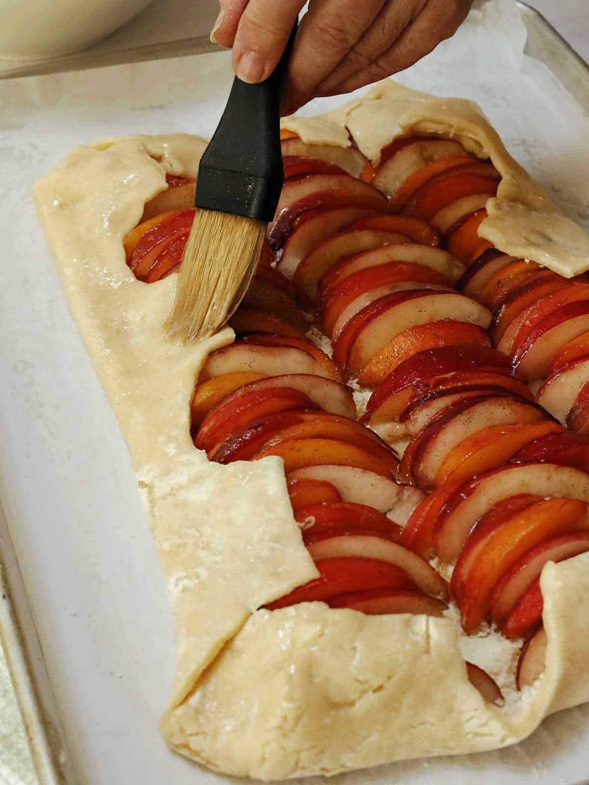 A hand uses a pastry brush to apply glaze to the edges of an unbaked fruit galette with sliced plums arranged on a baking tray.