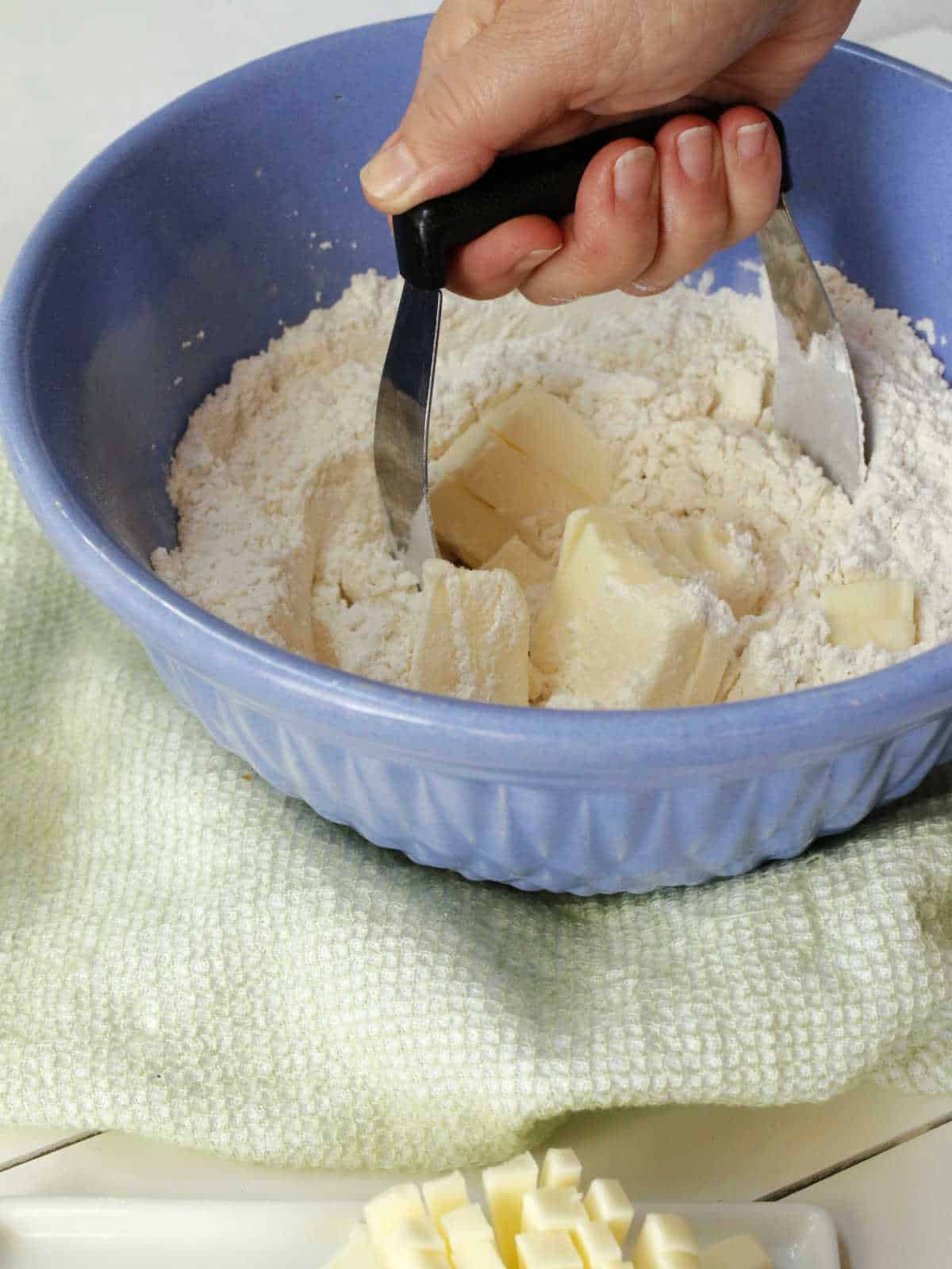 Hand using a pastry cutter to blend butter into flour in a blue bowl, with a green towel underneath and cubed butter nearby.