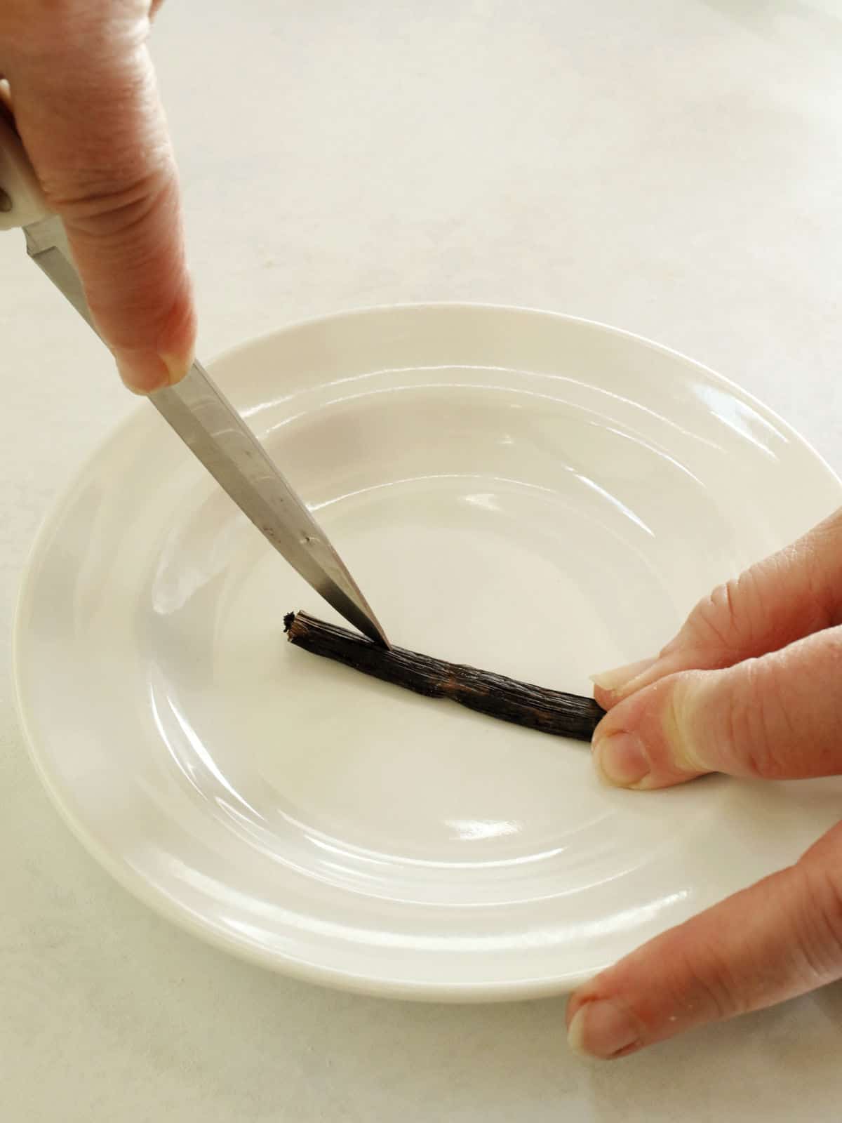 A person uses a knife to split open a vanilla bean pod on a white plate.