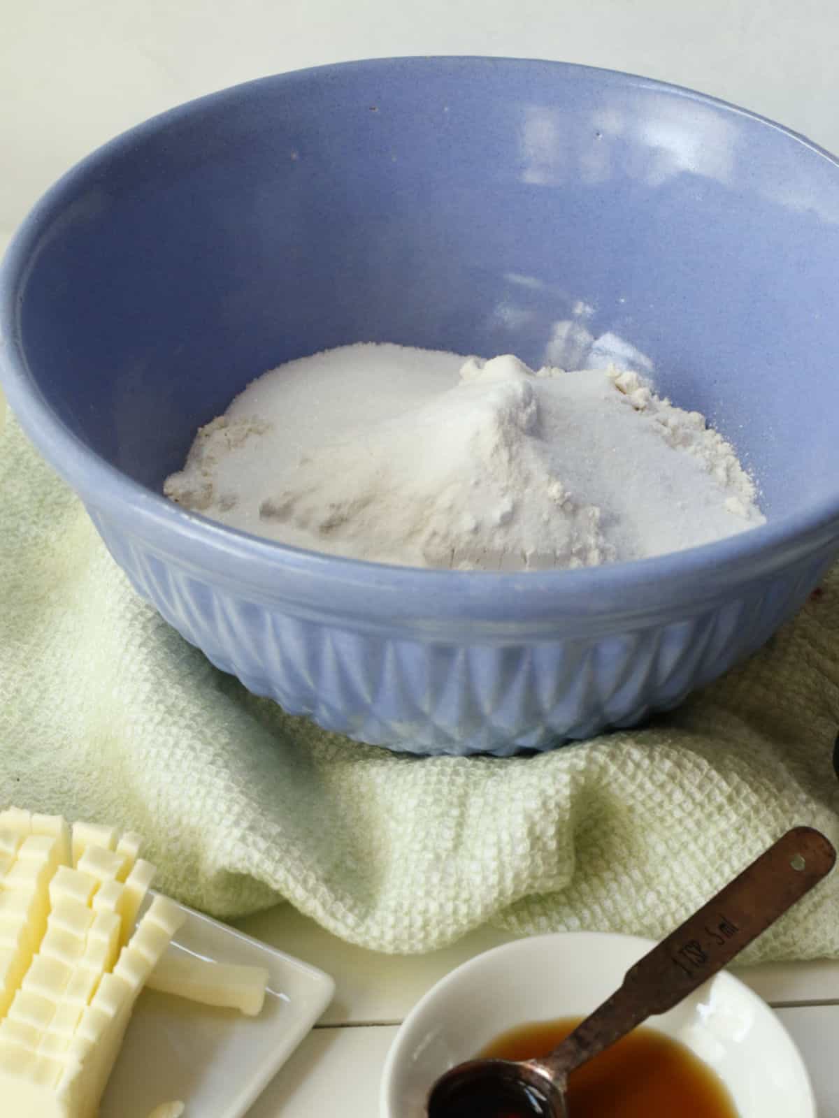 A blue bowl with flour and sugar sits on a green towel; butter slices, a tablespoon of vanilla, and a measuring spoon are nearby.