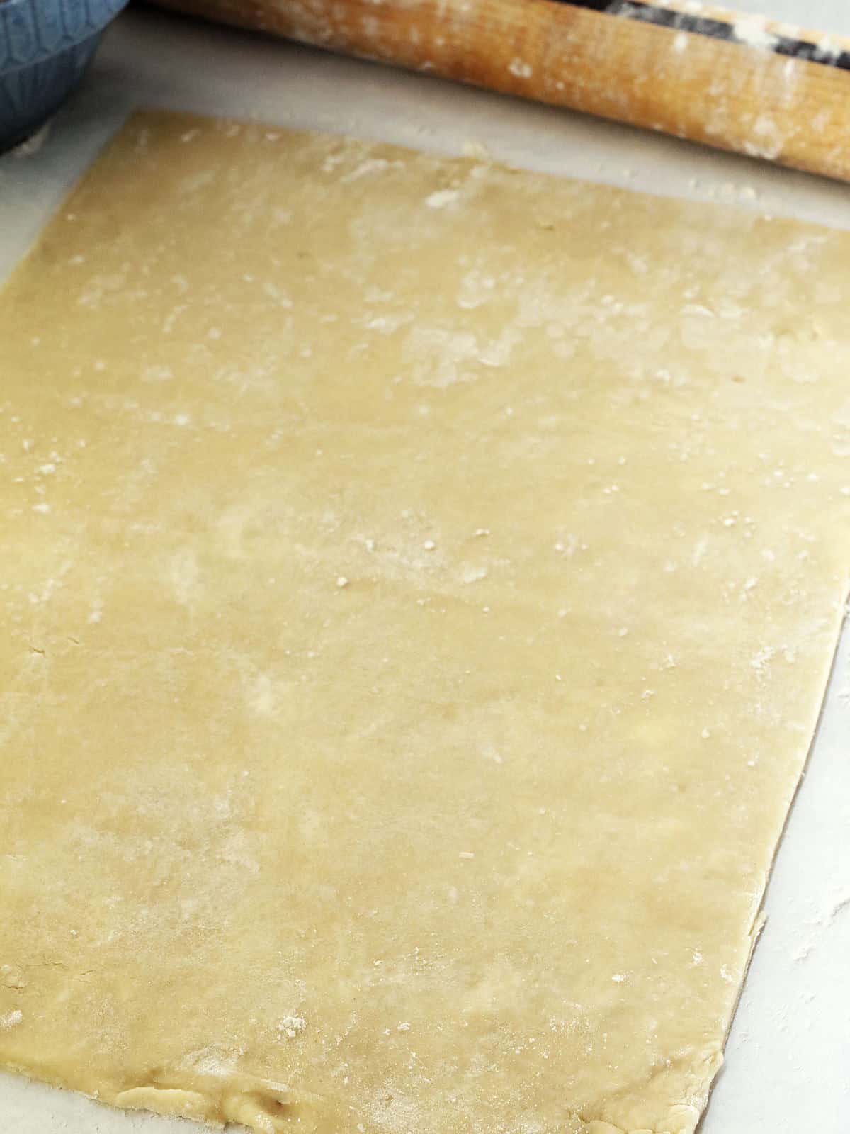A sheet of rolled-out dough on a floured surface with part of a rolling pin and a bowl visible at the edge of the frame.
