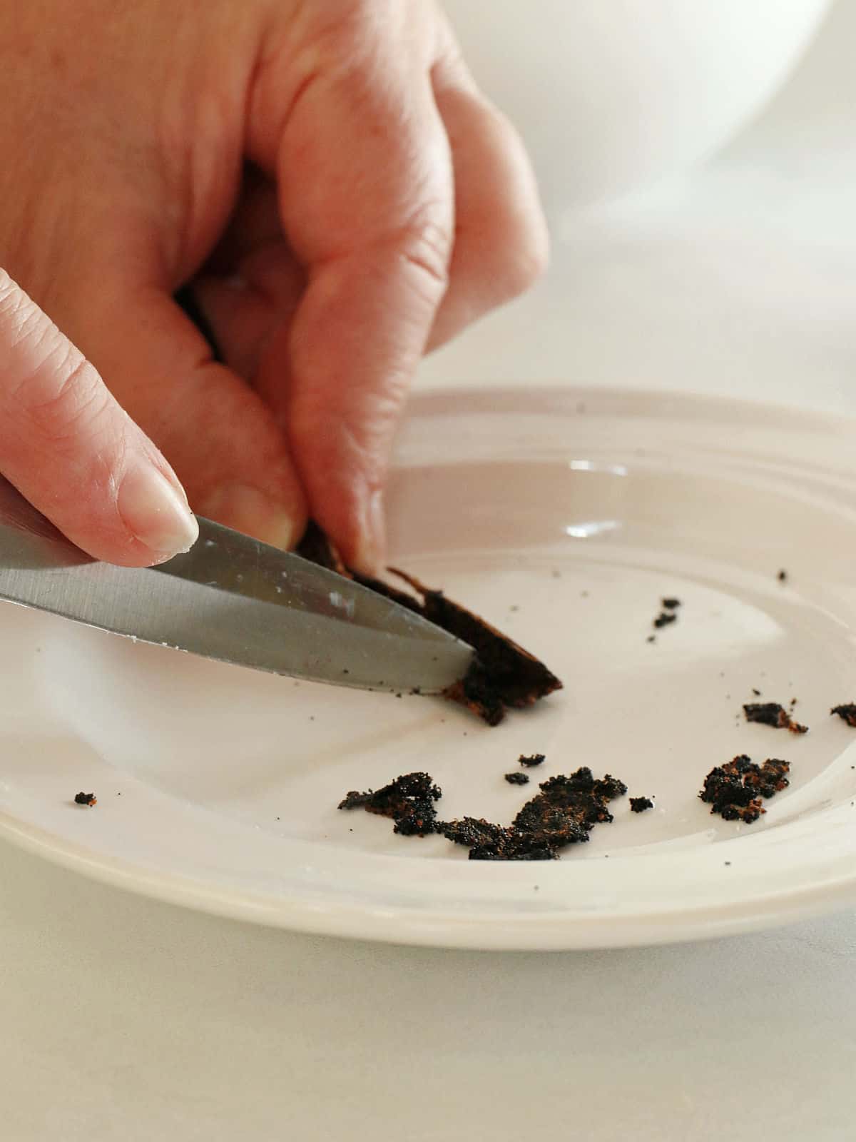 A hand uses a knife to scrape seeds from a vanilla bean pod onto a white plate.