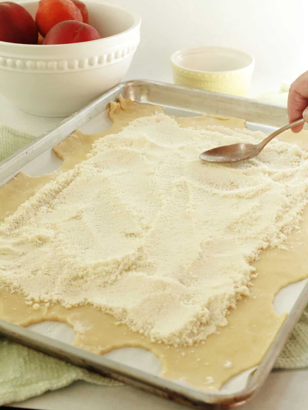 A hand spreads a layer of flour or sugar over rolled-out dough on a baking sheet. A bowl of peaches and a small cup are in the background.