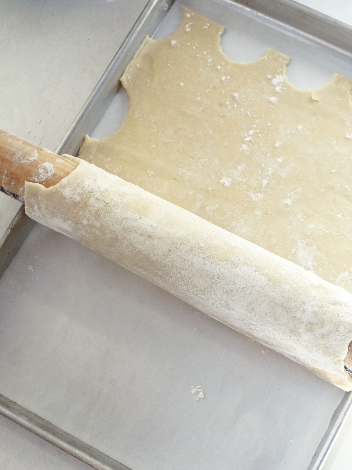 A rolling pin is rolling out dough onto a metal baking sheet lined with parchment paper.
