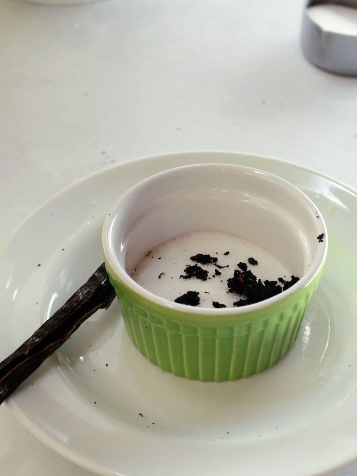A green ramekin with sugar and vanilla bean seeds sits on a white plate, with an empty vanilla pod next to it.