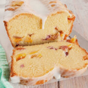 Iced peach loaf cake with two slices cut, showing chunks of peaches inside, on a light wooden surface next to a green striped cloth.